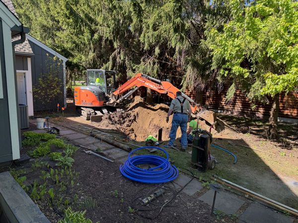 Orange mini excavator digging a deep trench in a backyard while two workers stand nearby with blue coiled pipe and equipment.