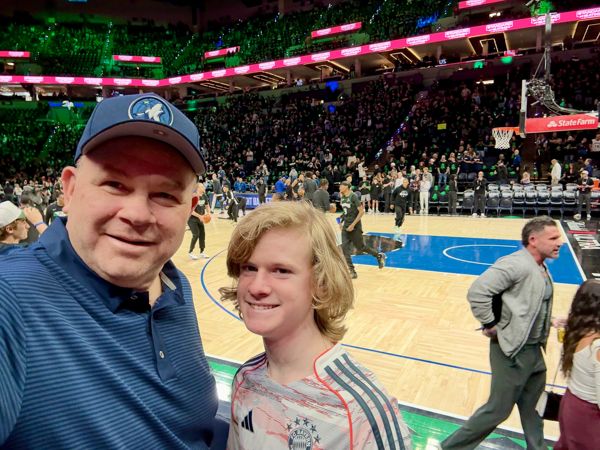 Man in blue Timberwolves cap and boy with shaggy blond hair smiling courtside at a packed NBA arena during warmups.