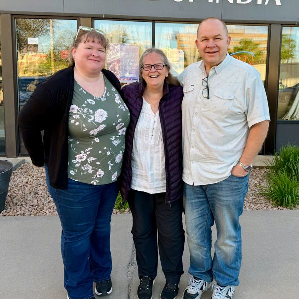Three people smiling arm-in-arm outside an Indian restaurant on a sunny day.