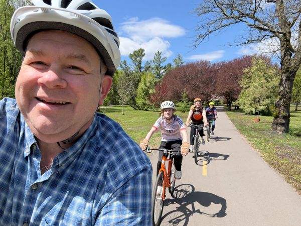 Man in white helmet takes a selfie while three cyclists follow behind on a sunny paved trail through a park.