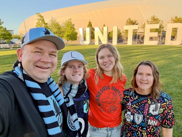 Four people taking a selfie in front of a large UNITED sign and stadium, wearing Minnesota United scarves and shirts.