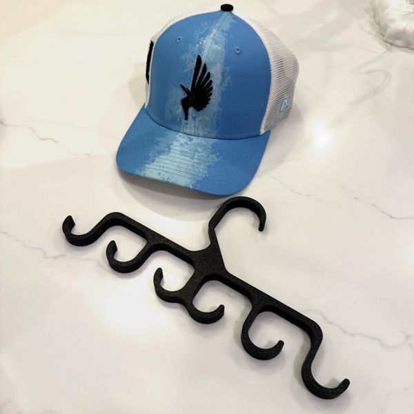 Blue and white trucker cap with a bird logo beside a black 3D-printed hat hanger with multiple hooks, on a marble surface.