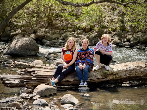 Three people sitting on a fallen log in a rocky creek, smiling at the camera, surrounded by trees and flowing water.