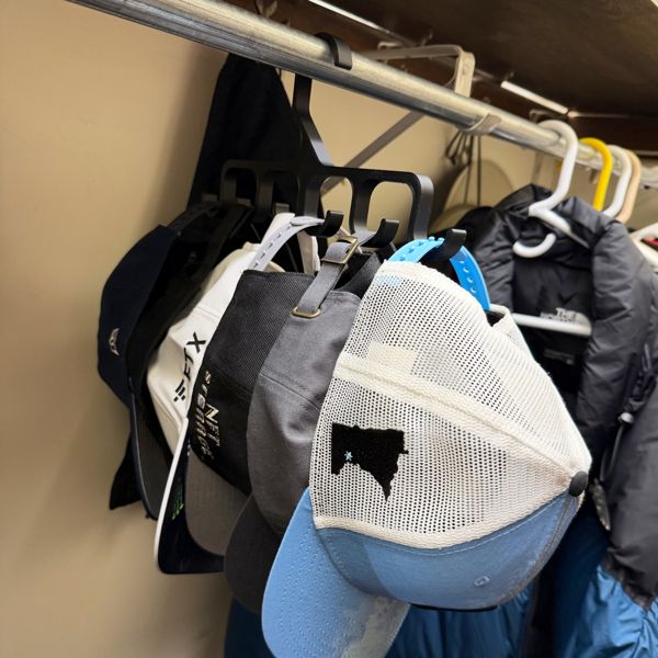 Six baseball caps hanging from a 3D-printed black hat hanger hooked over a closet rod, with jackets visible behind them.