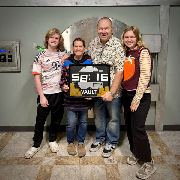 Four people posing in front of a large vault door holding a sign showing their escape room completion time of 58 minutes 16 seconds.