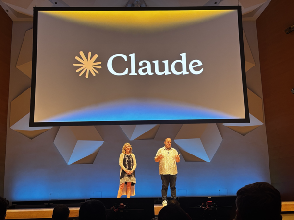 Two people standing on stage in an auditorium presenting in front of a large screen displaying the Claude logo and wordmark.
