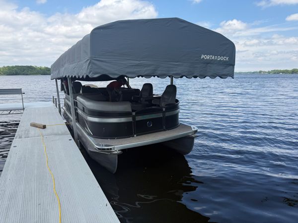 Pontoon boat with gray Porta-Dock canopy cover docked at a floating dock on a calm lake under partly cloudy skies.
