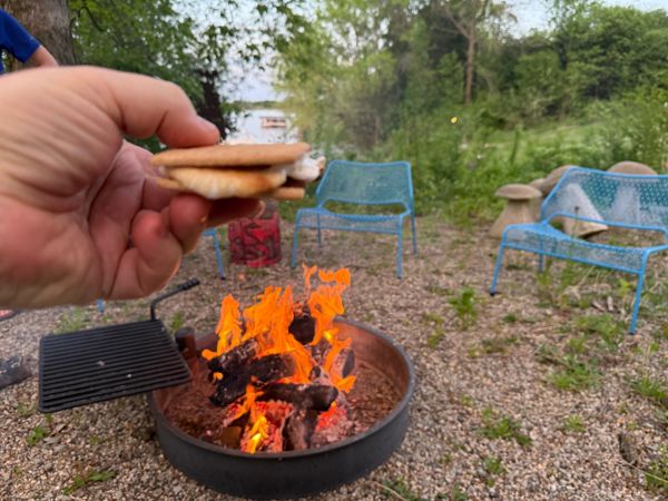 Hand holding a s'more over a campfire in a metal fire pit, with blue metal chairs and trees in the background.