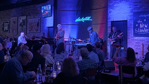 Live band performing at Dakota jazz club with audience seated at tables in dim blue and red stage lighting.