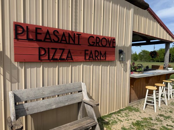 Exterior of Pleasant Grove Pizza Farm building with red sign, a wooden bench, and a covered bar area with stools.