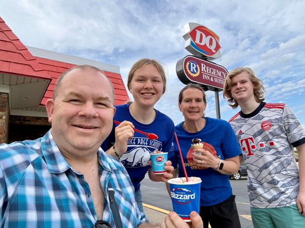 Four people smiling and holding Dairy Queen Blizzard cups in front of a DQ sign on a sunny day.