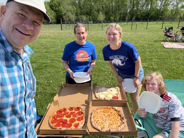 Four people smiling outdoors at a picnic table holding plates over three open pizza boxes on a sunny grassy field.