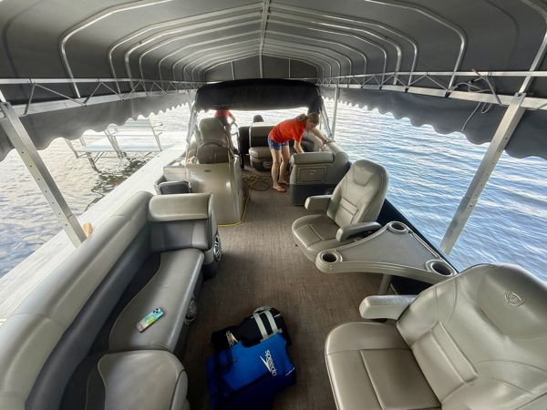 Two people cleaning a large double-decker pontoon boat with gray leather seating and a canopy top on calm water.