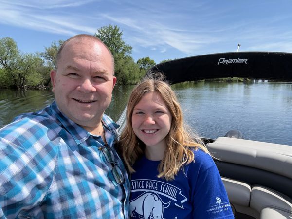 Man and teenage girl smiling together on a Premier pontoon boat on a calm river, trees in background on a sunny day.