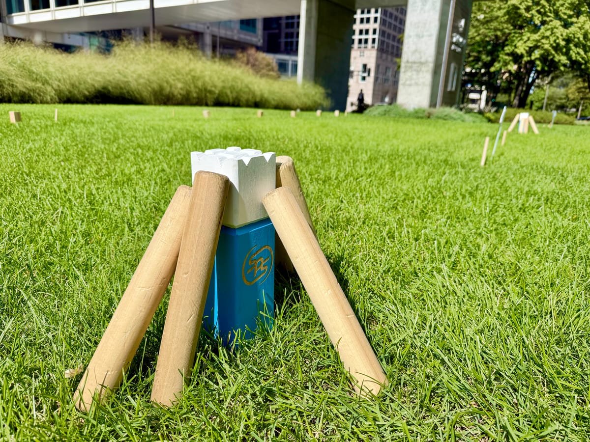 Kubb game in progress on a sunny lawn with wooden batons leaning against a blue and white king piece
