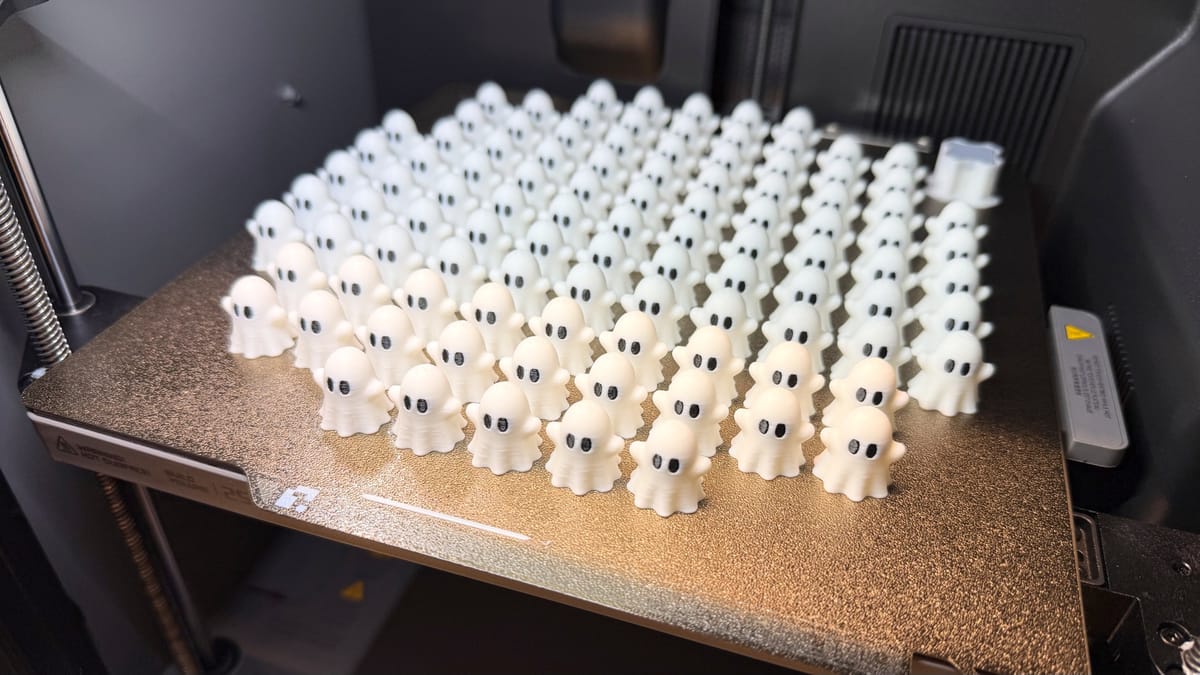 Rows of printed white ghost figures with black eyes standing on a textured metal build plate inside a 3D printer.