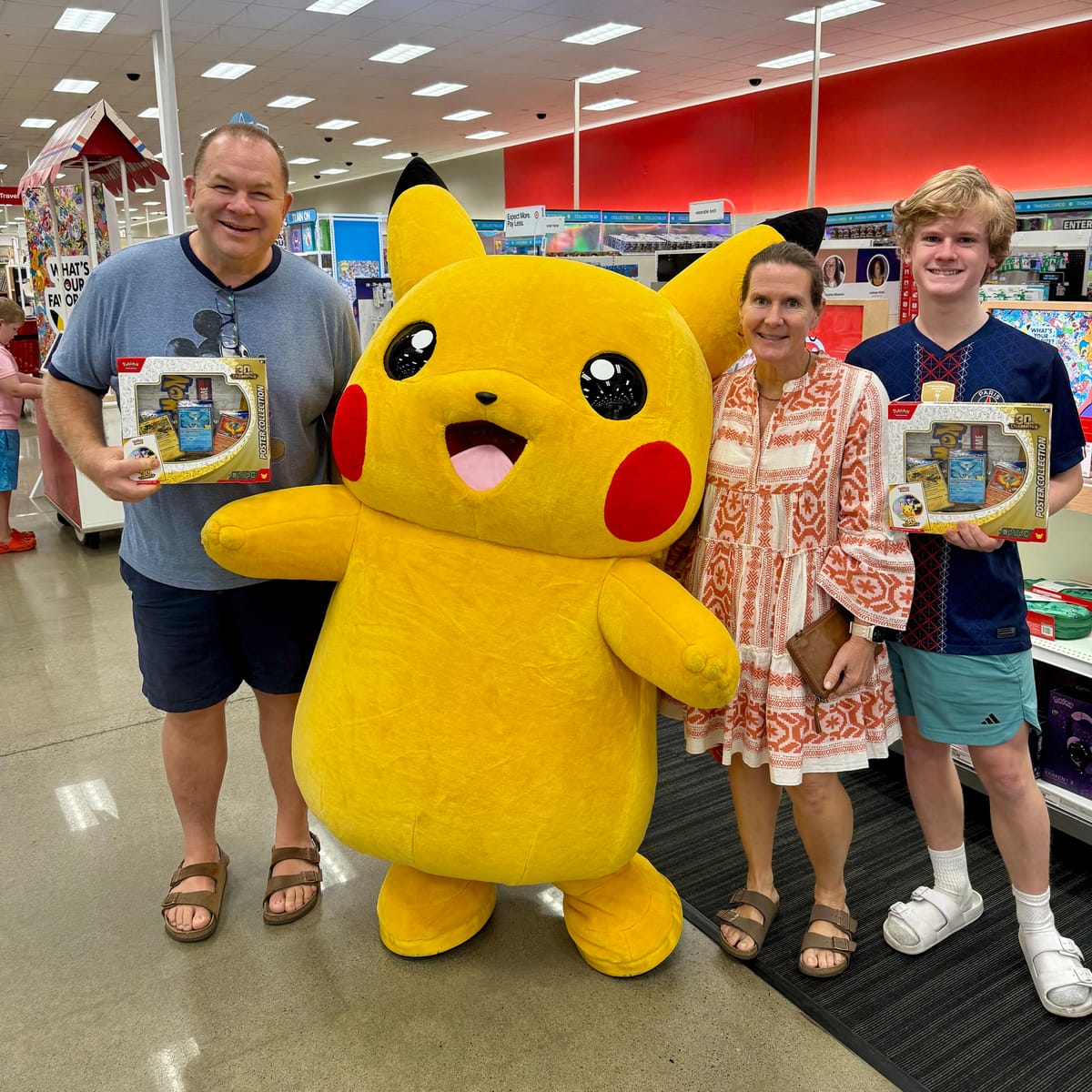 Jamie, Tammy, and Tyler pose in a store aisle with a costumed Pikachu, two holding boxed Pokémon card sets.