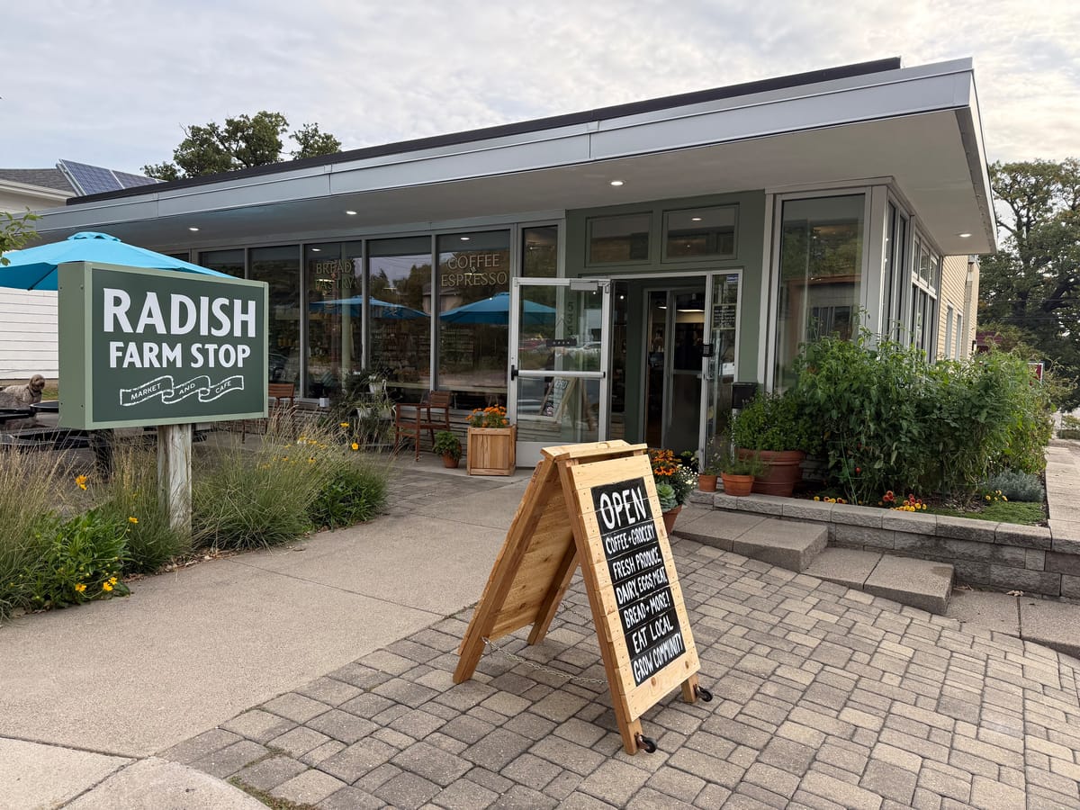 Glass-fronted Radish Farm Stop market and cafe with a green sign, a wooden sandwich board reading OPEN, and planters by the door.