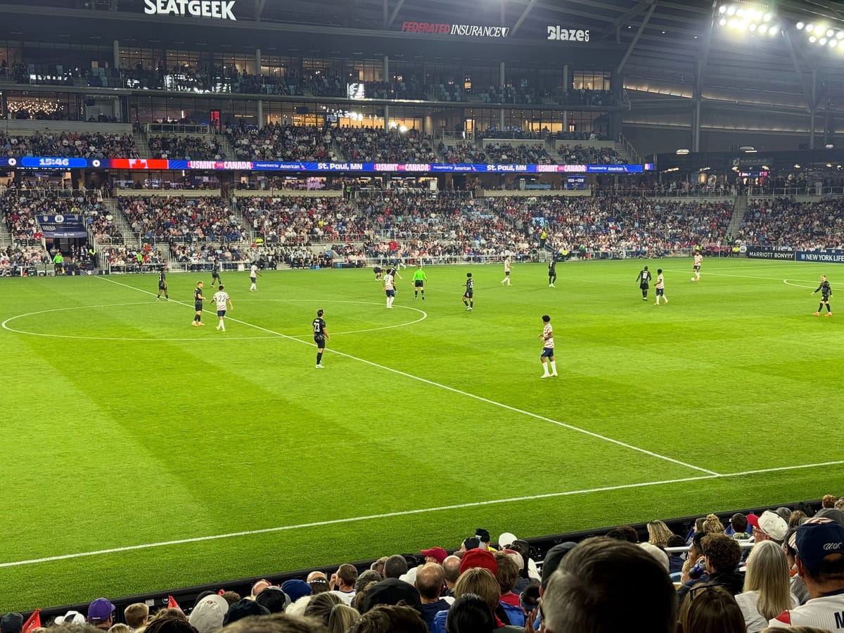 Players in white and black kits spread across a lit soccer pitch near midfield, with full stands and a ribbon board behind.