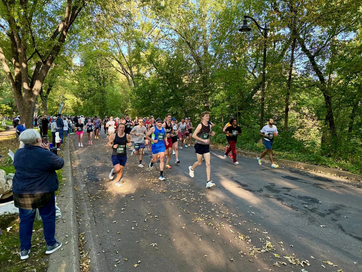 Runners wearing race bibs move along a tree-lined road scattered with leaves while spectators watch from the curb.