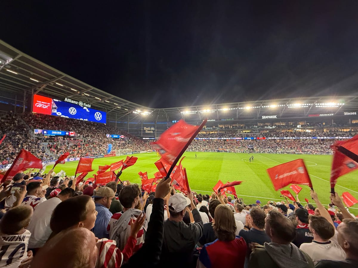 Fans in the lower stands wave red flags at night while the stadium video board shows a US goal graphic.