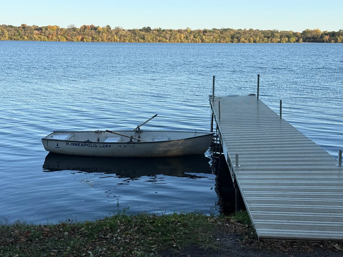 A white rowboat marked Minneapolis Lakes floats beside an aluminum dock, oars resting across its seats, on calm blue water.