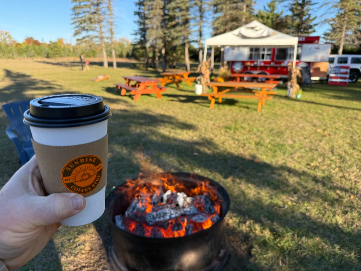 Hand holding a Sunrise Coffee Stop cup beside a glowing fire pit, with picnic tables and a red food trailer on the lawn