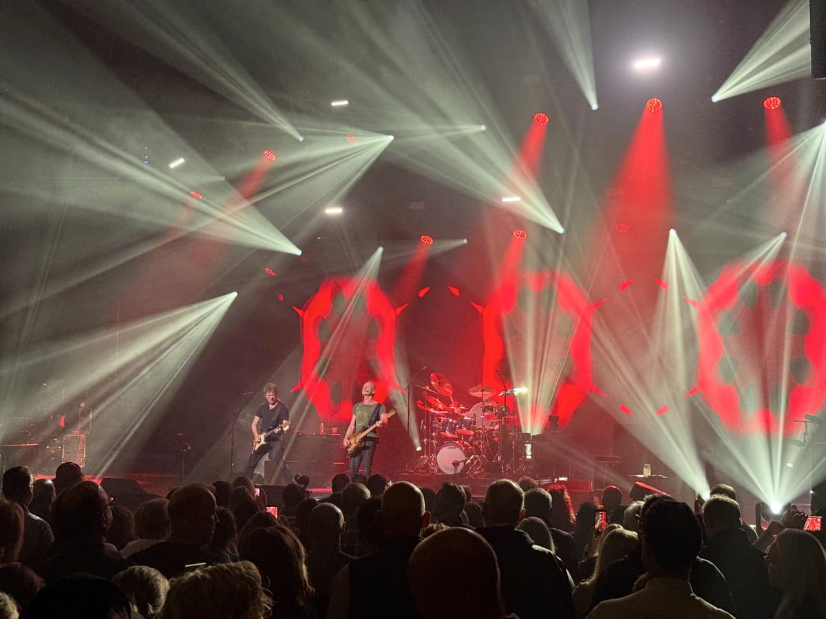 Guitarist, bassist, and drummer perform on stage under white light beams and red patterned projections above a crowd.