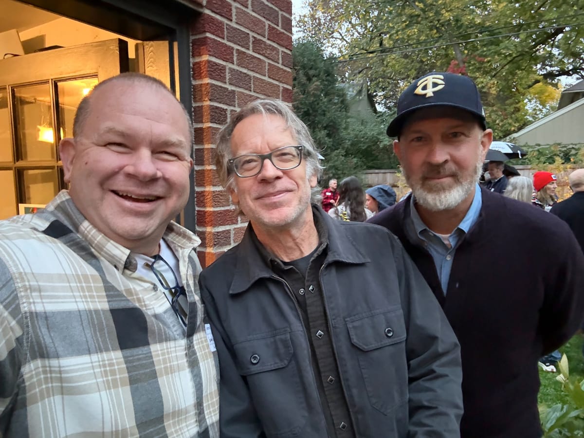 Three men pose for a selfie beside a brick house at a backyard gathering; one wears glasses, another a Twins cap.