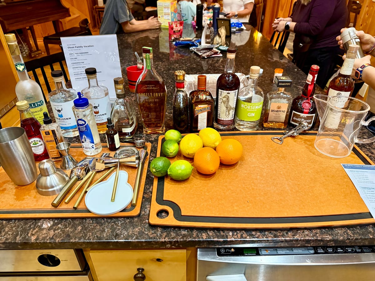 Home cocktail bar on a granite counter with liquor bottles, citrus fruit, shakers, jiggers, and a printed drink menu