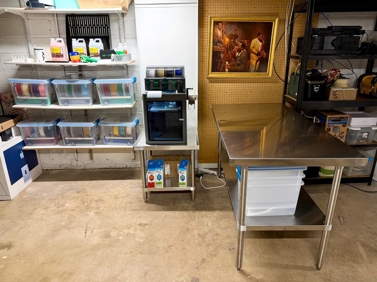 Basement corner with a 3D printer on a steel stand, shelves of filament spool bins, a steel worktable, and a jazz painting.