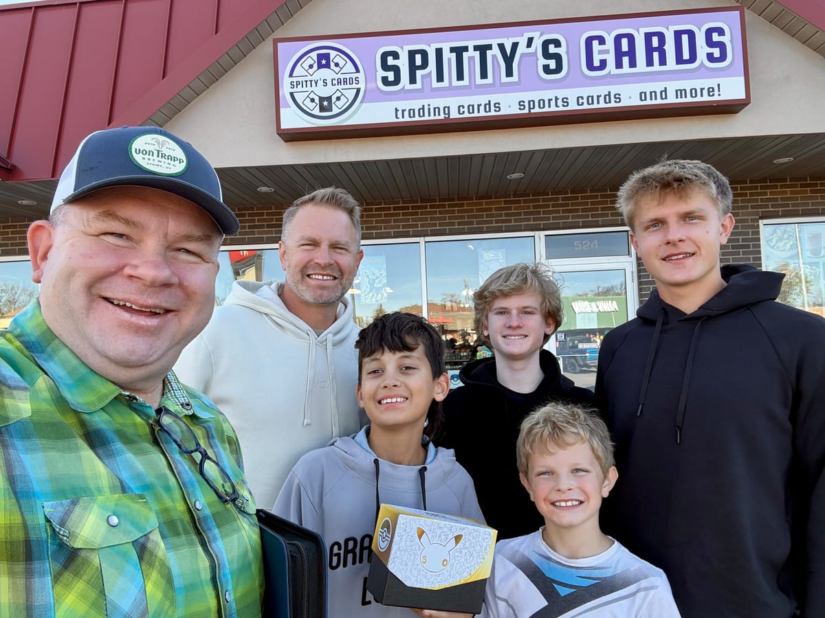 Six people smile outside Spitty's Cards, a boy in front holding a Pokémon card box