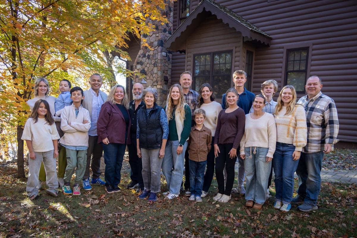 About twenty adults and children of several ages pose on leaf-covered grass beside a brown log house and yellow-leafed tree.