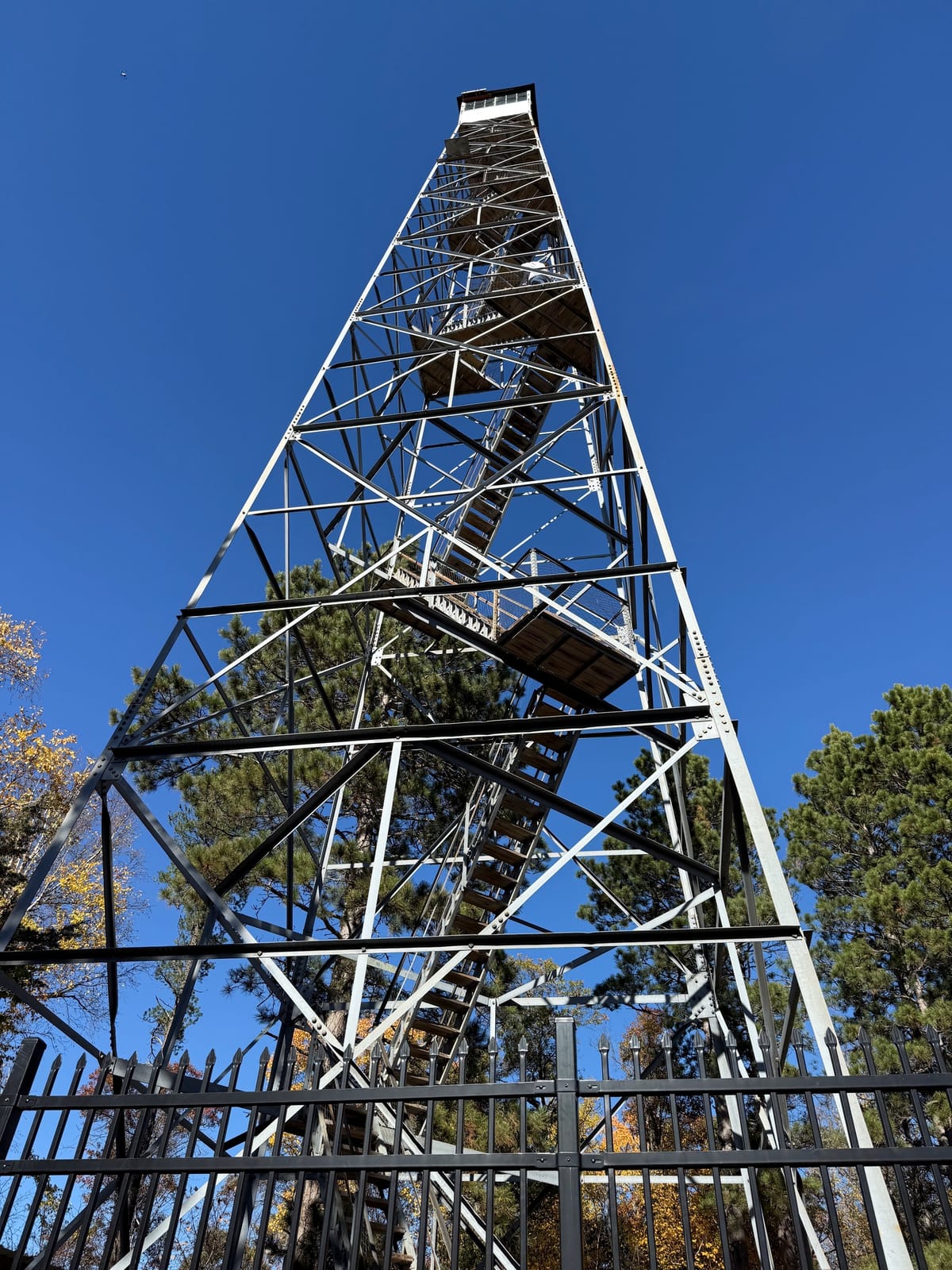 Steel lattice tower with switchback wooden stairs rising to a small cab, seen from below behind a black iron fence and pines.