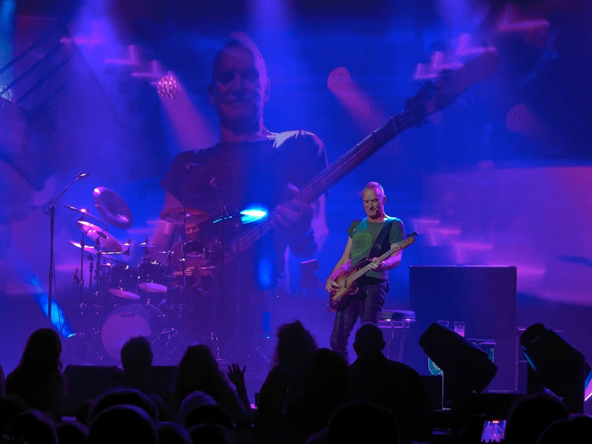 Sting plays bass under purple light beside a drummer, a giant screen of him behind them and audience silhouettes below.